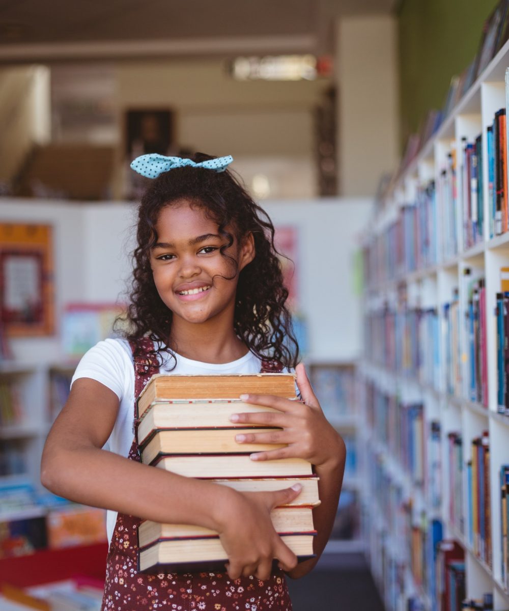 portrait-of-smiling-african-american-schoolgirl-carrying-stack-of-books-in-school-library.jpg